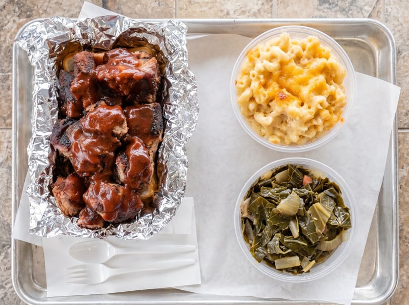 BBQ ribs in foil with mac and cheese and collard greens in plastic containers on a tray.