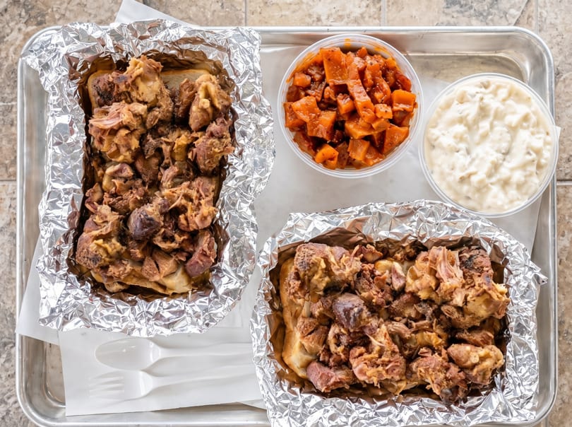 Two foil-wrapped sandwiches with meat, a side of sweet potatoes, and a side of potato salad.