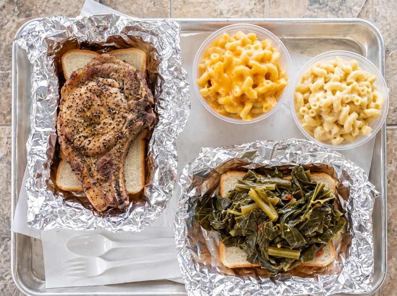 A takeout tray with a pork chop, collard greens, and two containers of macaroni and cheese.