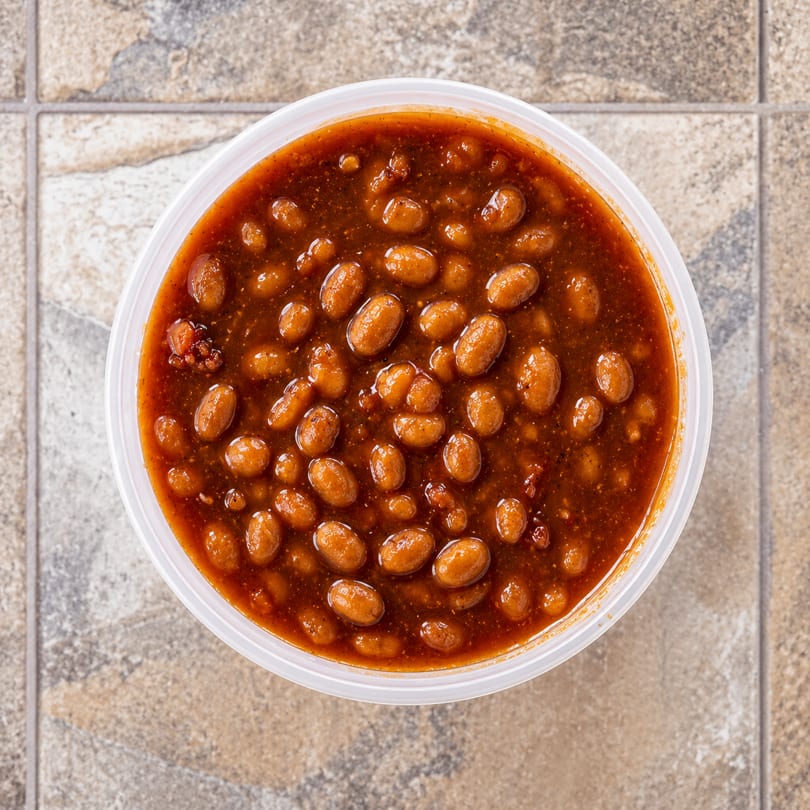 Overhead view of a white plastic container filled with baked beans in a rich, dark sauce.
