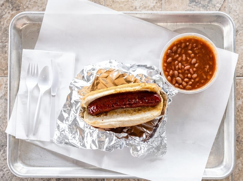 A hot dog in a bun wrapped in foil, with a side of baked beans and plastic cutlery.